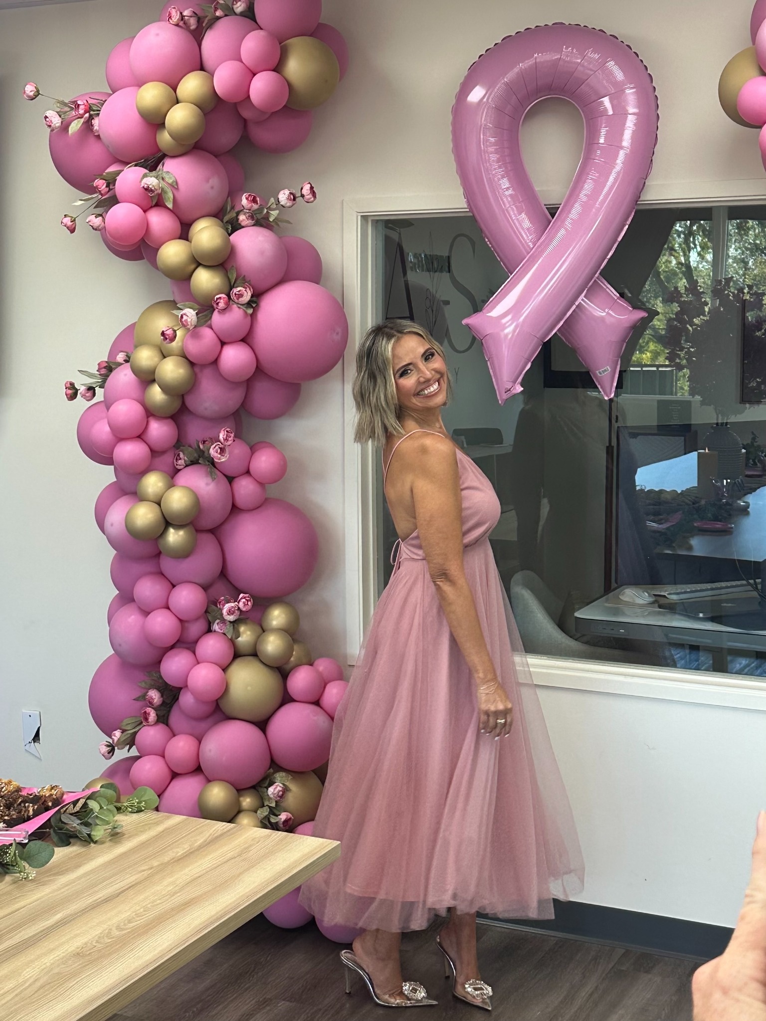 Celeste in her pink dress beside pink and gold balloons and a breast cancer ribbon